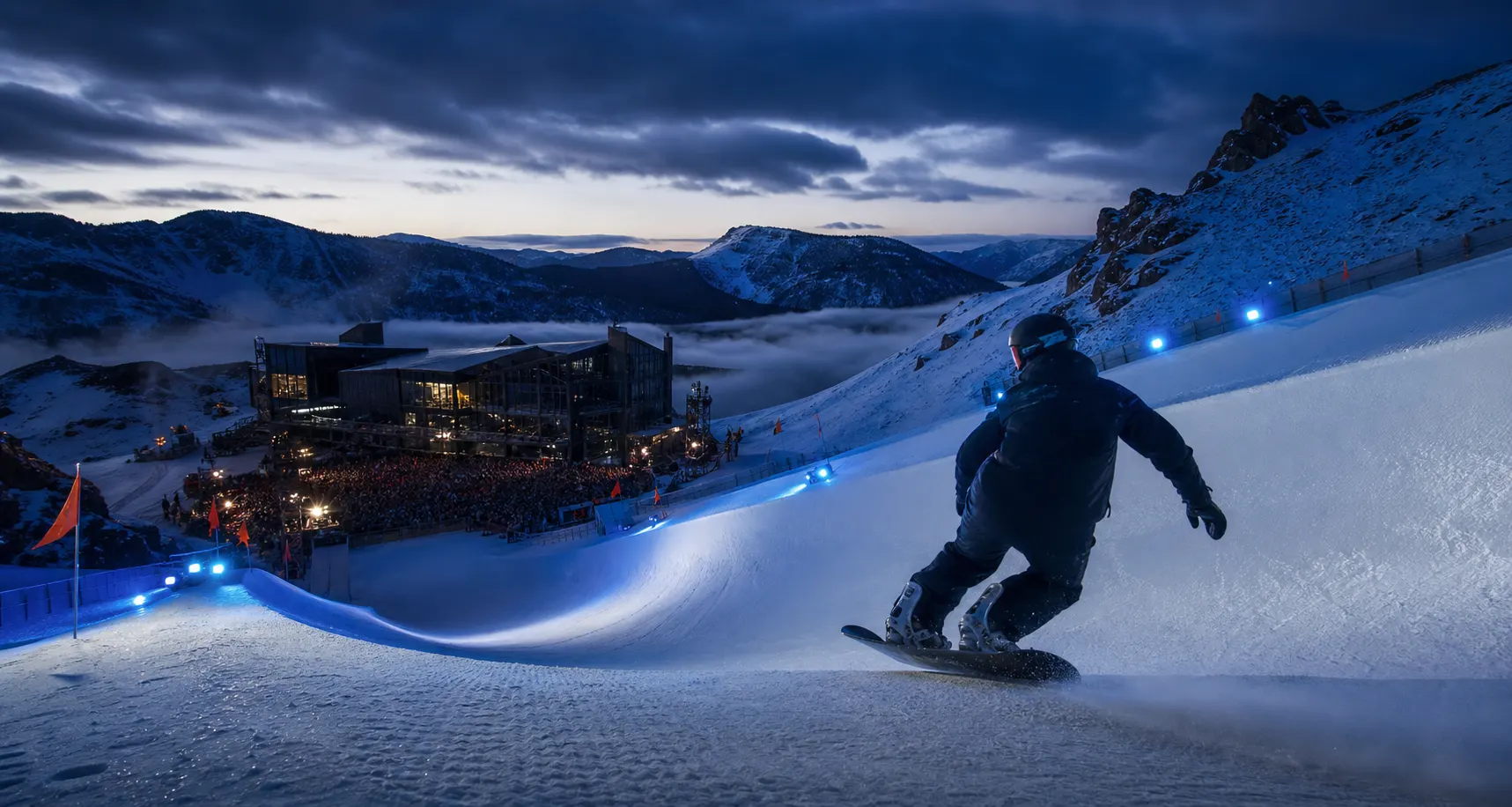 A snowboarder descends an illuminated course toward a mountain music hall at blue hour.